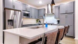 Una cocina moderna con gabinetes grises, electrodomésticos de acero inoxidable, una isla central con taburetes altos e iluminación colgante.