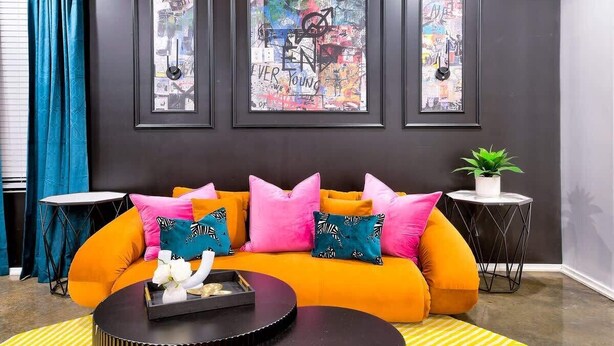 Una sala de estar moderna con un sofá naranja brillante, varios cojines coloridos y tres cuadros en la pared.