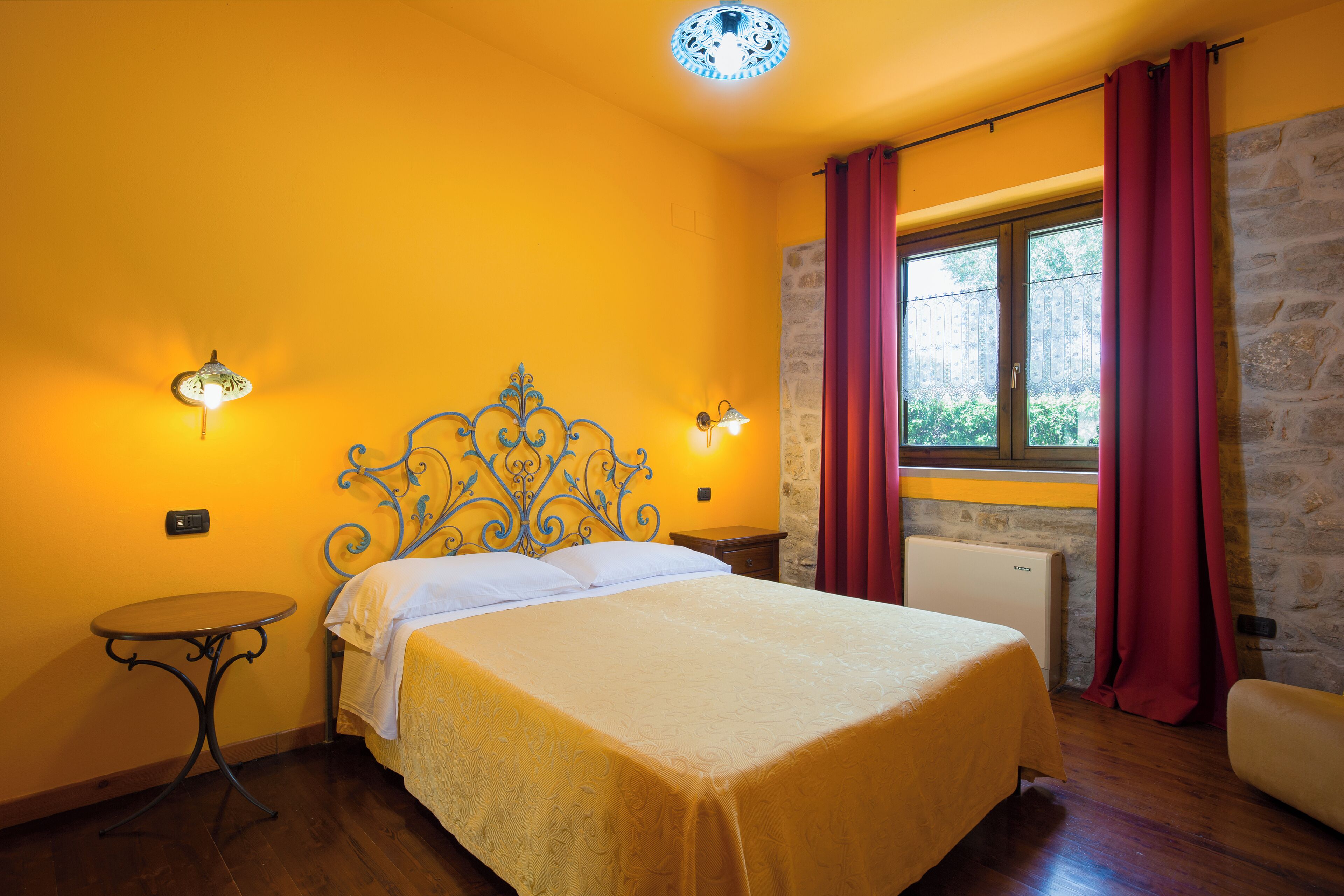 A bedroom with a bed, a small table, a nightstand, a stone wall, a window with curtains, and a ceiling light fixture.