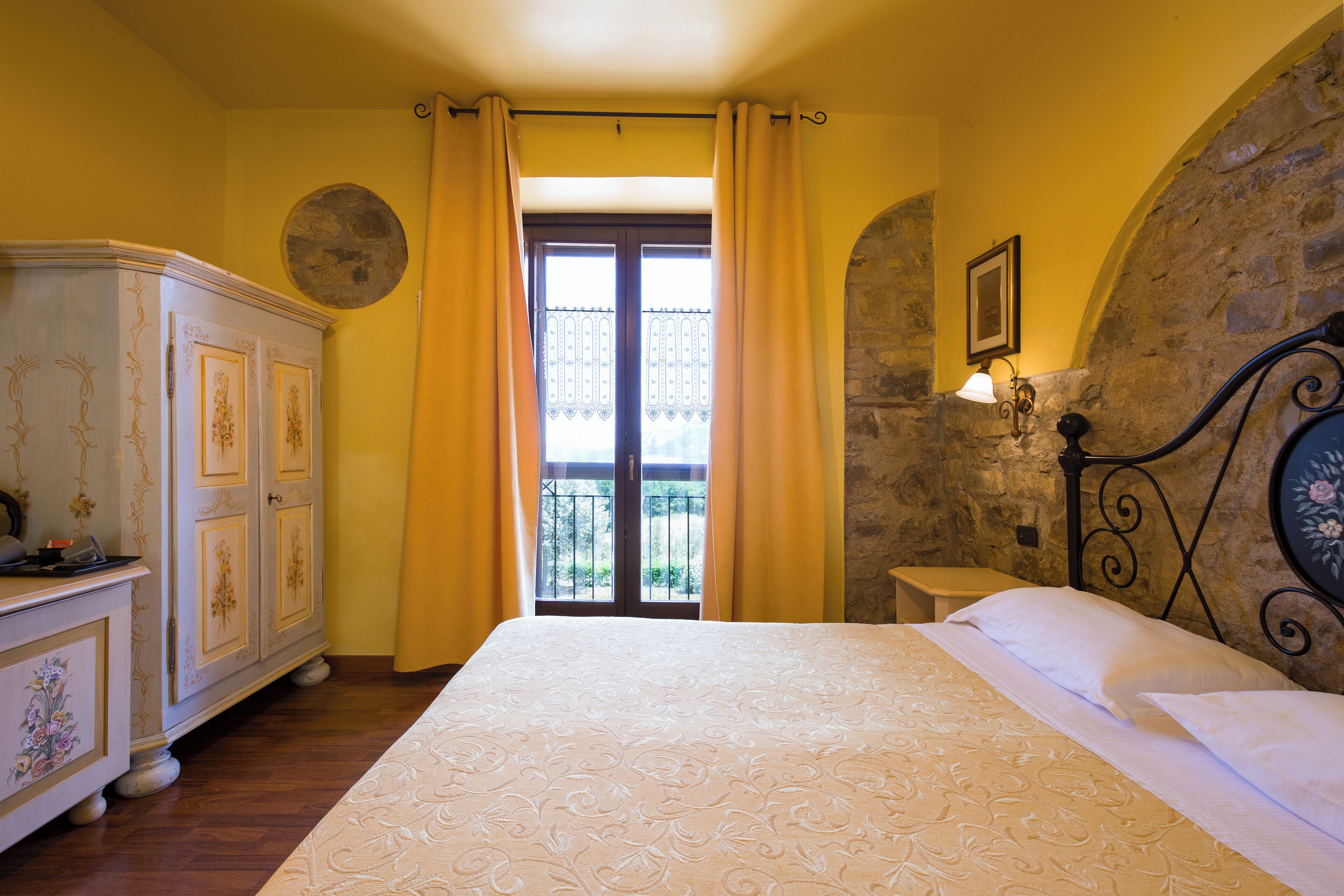 Une chambre à coucher avec un lit, une commode, une fenêtre avec des rideaux, un mur en pierre et une petite table.