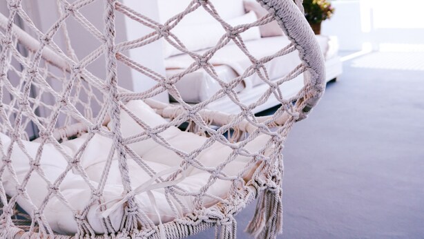 Una cadira penjant de macramé amb un coixí blanc, suspesa per cordes, davant d'una paret blava.