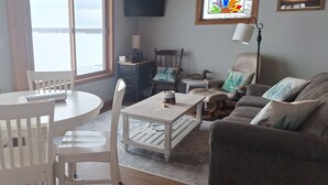 Een gezellige woonkamer met een ronde tafel, witte stoelen, een grijze bank en een salontafel. Er is een raam met uitzicht op een wateroppervlak.