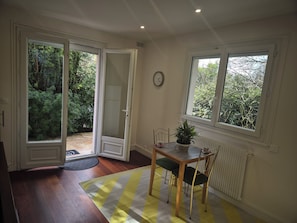 A room with a dining table, chairs, a potted plant, and a view of greenery through the windows. - Renovated one-bedroom apartment in a quiet neighborhood with a flower-filled terrace (Le Raincy)