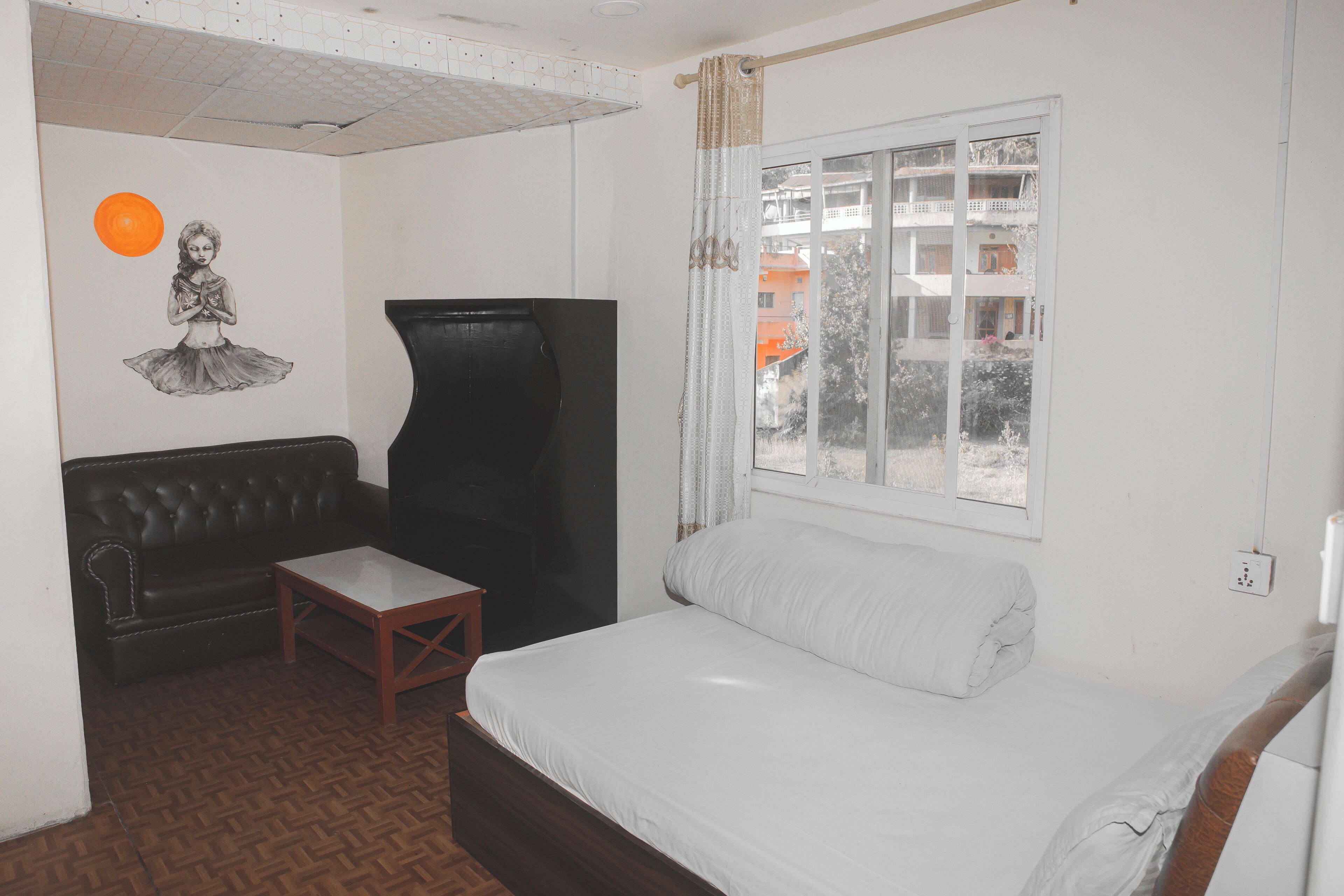 A bedroom with a bed, a black leather sofa, a small wooden table, a black TV stand, and a wall with a drawing.