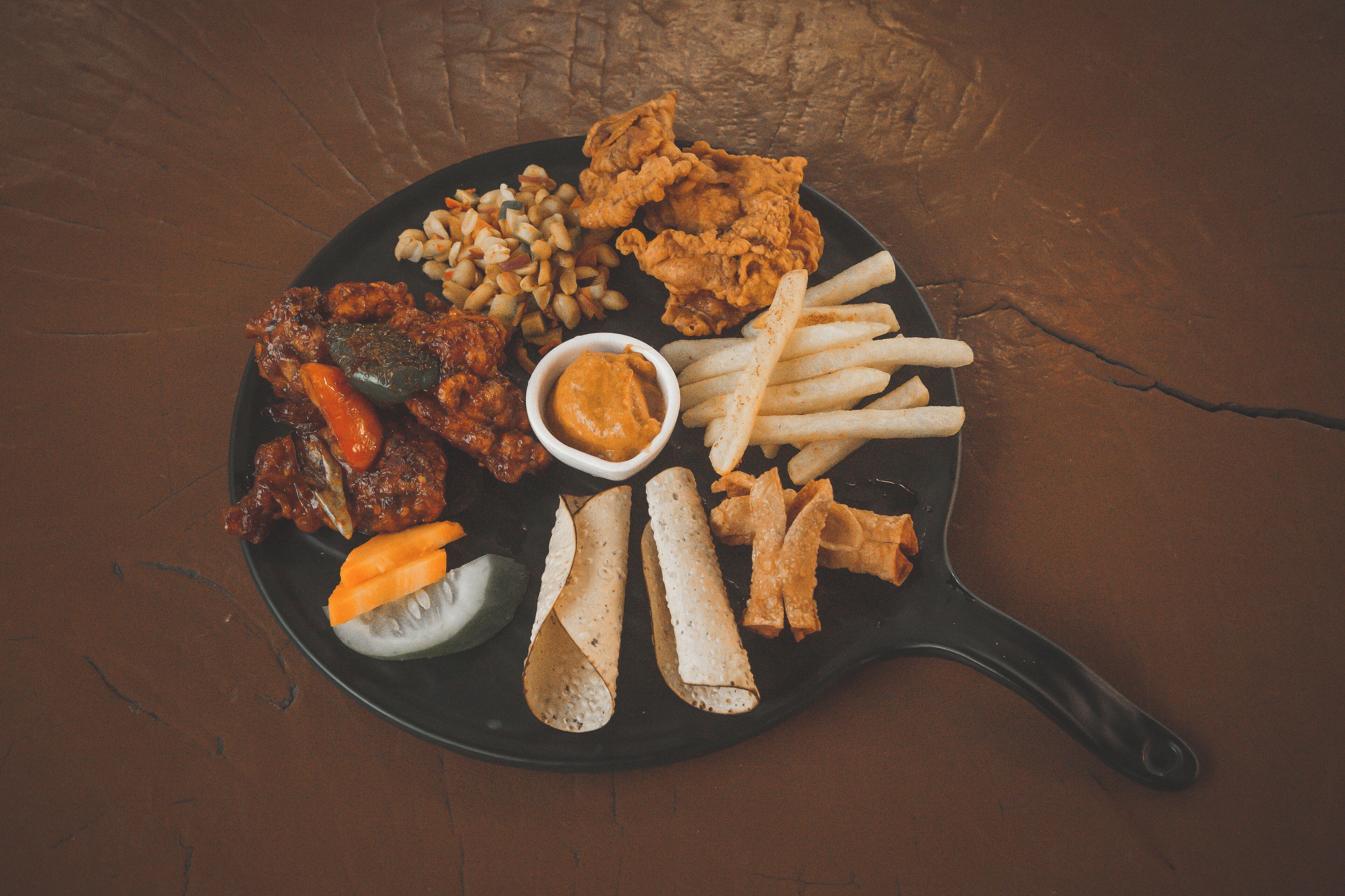 A black plate with fried chicken, French fries, a small bowl of dipping sauce, and some vegetables.