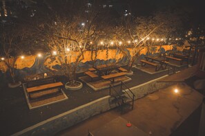 An outdoor seating area with benches and tables, surrounded by trees and illuminated by string lights. - Gypsy hostel & Backpackers (Pokhara)