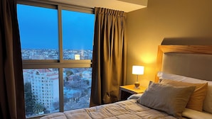 Une chambre d’hôtel dotée d’une grande fenêtre, d’un lit avec une tête de lit matelassée, d’une table de chevet avec une lampe et offrant une vue sur le paysage urbain.