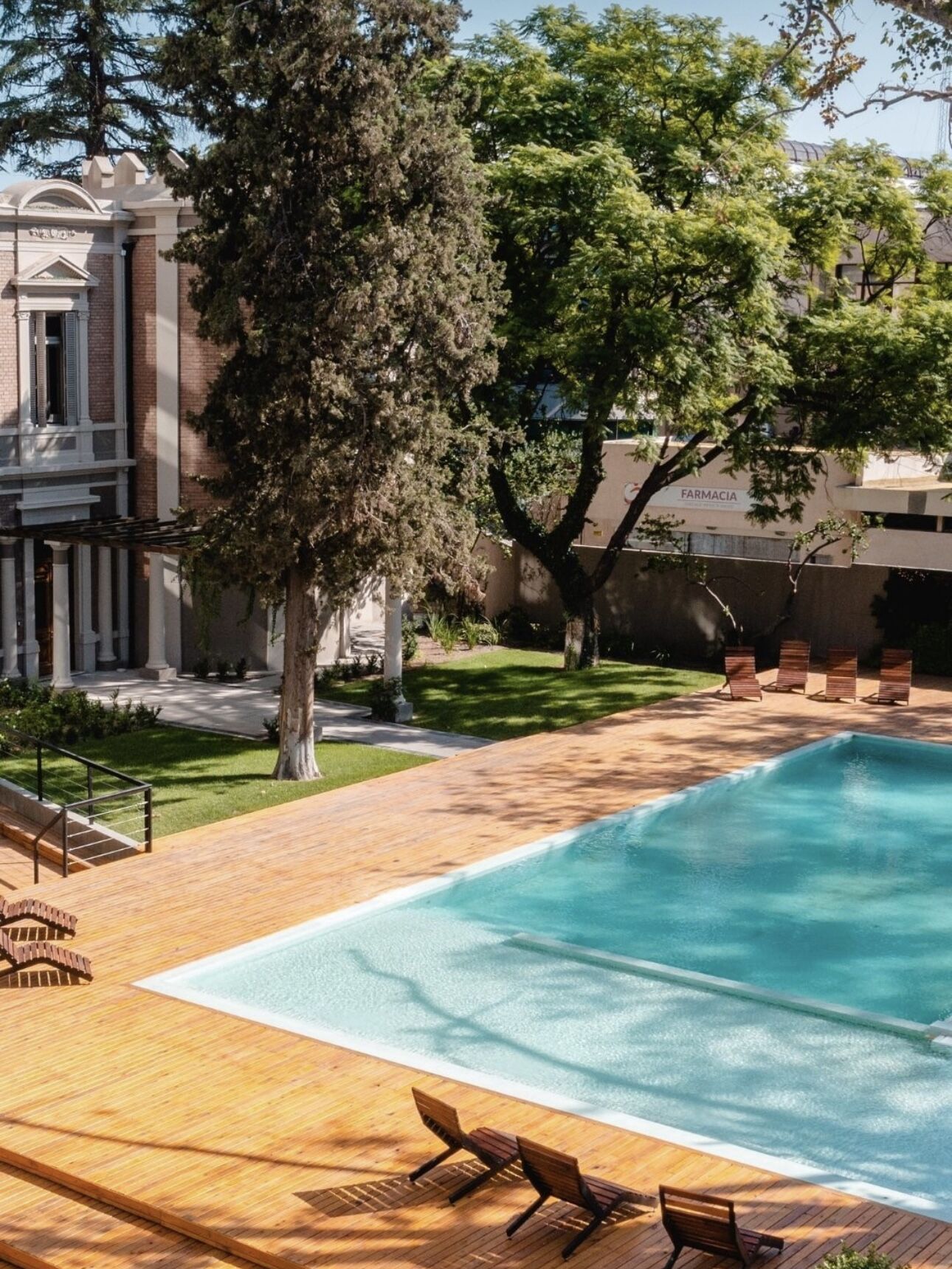 A swimming pool with wooden deck and lounge chairs, surrounded by trees and a building labeled "FARMACIA".