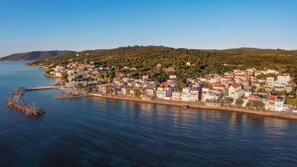 A coastal town with buildings along the waterfront, a rocky breakwater, and hills in the background.