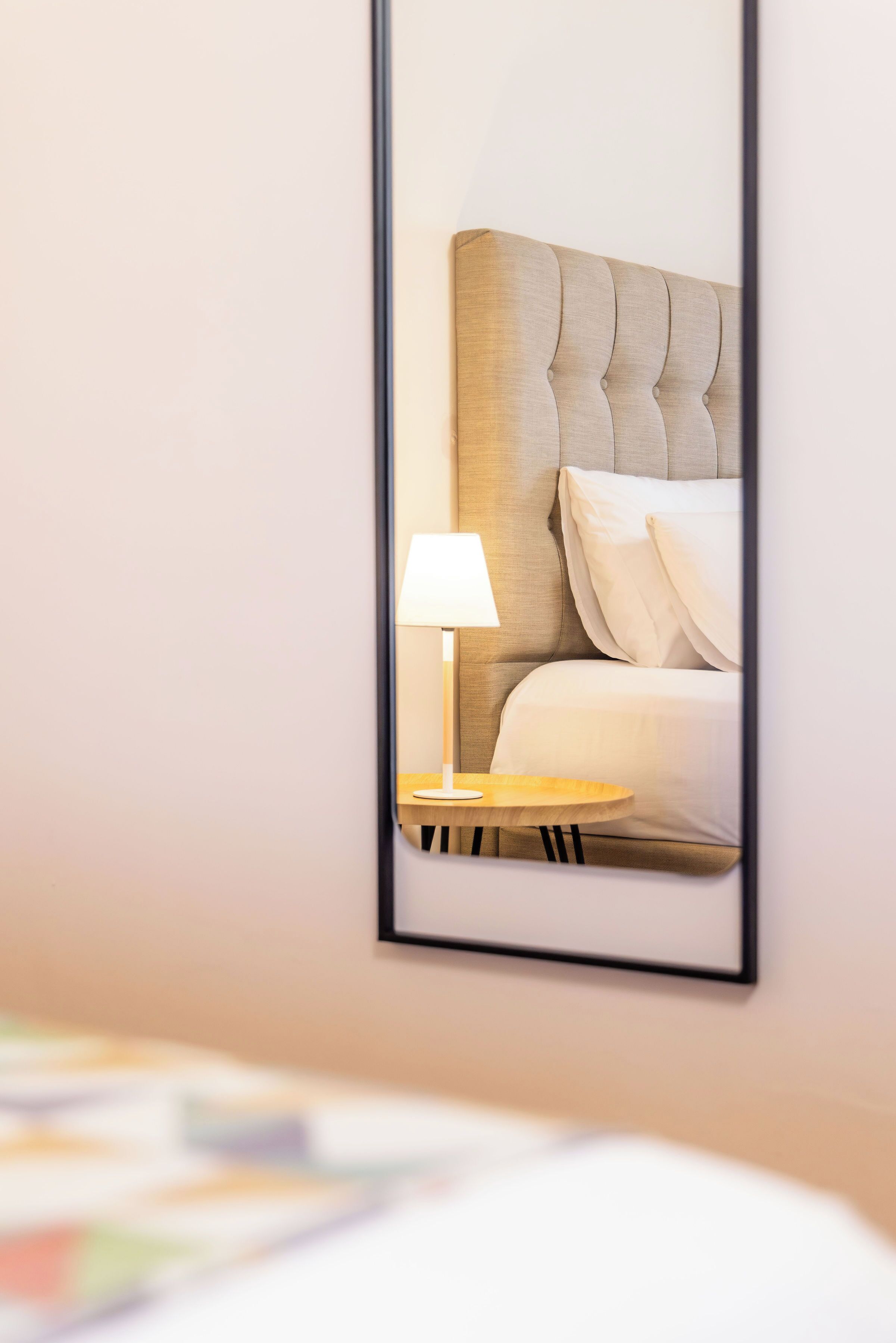 A framed mirror reflecting a bed with a tufted headboard, a bedside lamp, and a round wooden nightstand.