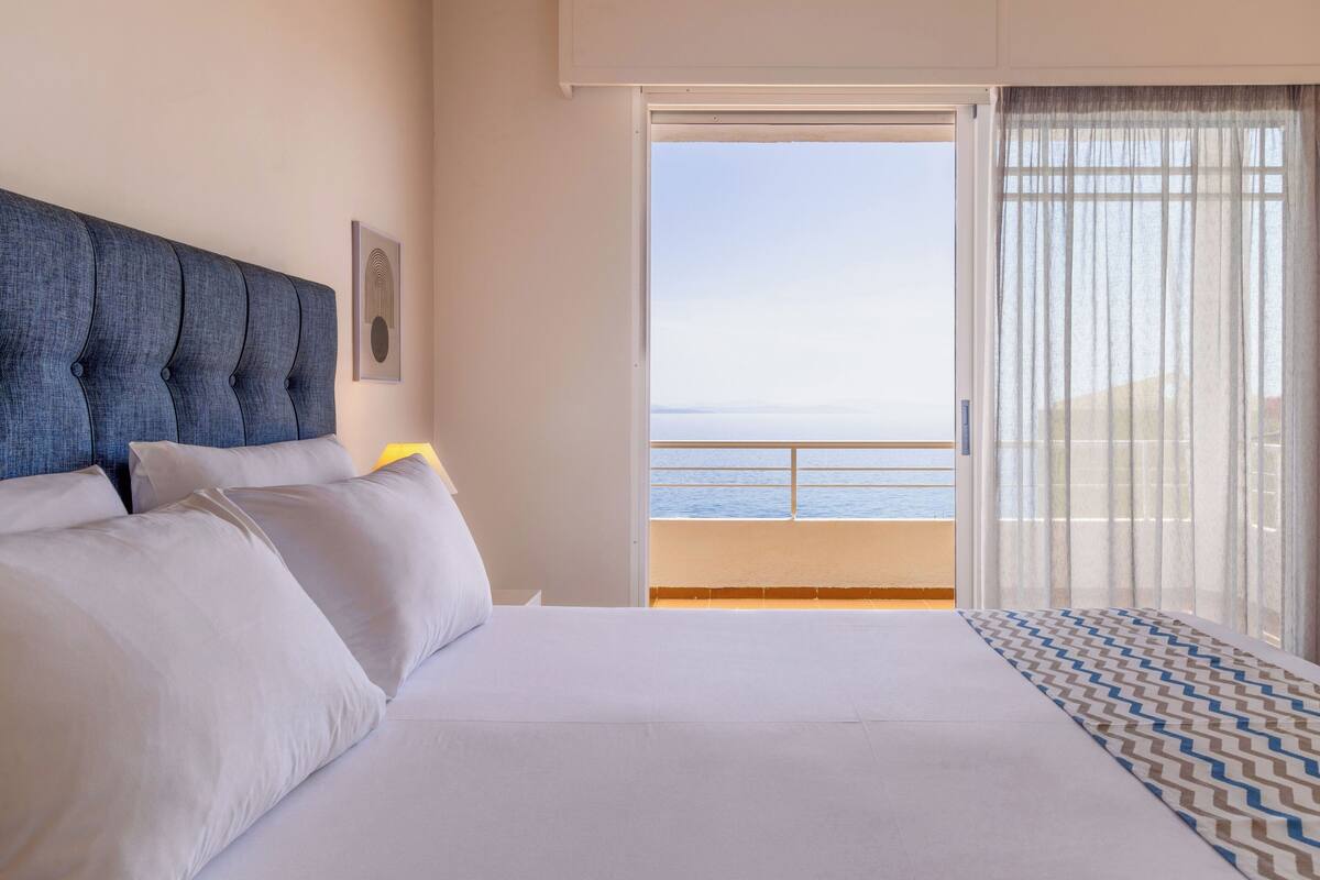 Habitación de hotel con una cama, cabecero, ventana con vista al mar y alfombra estampada.