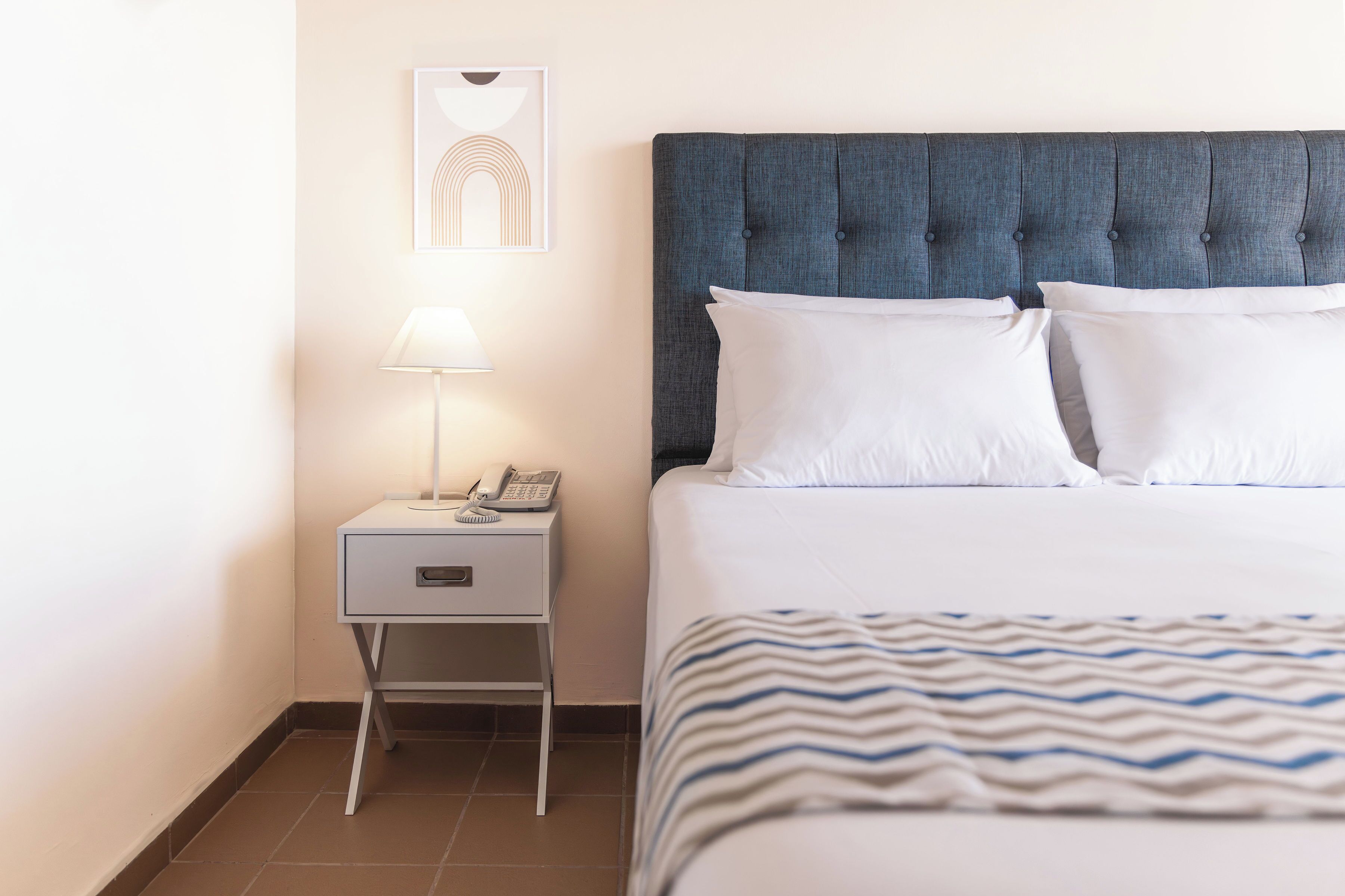 A neatly made bed with white pillows and a blue headboard, a bedside table with a lamp and a phone, and a framed picture on the wall.