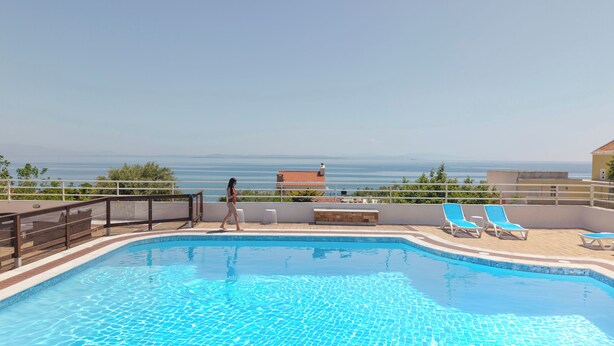 A swimming pool with a view of the sea, a person walking, and lounge chairs.