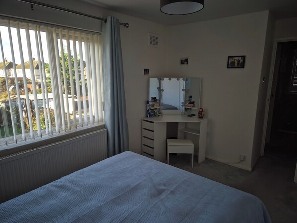 A bedroom with a bed, a vanity with a mirror, and a window with blinds.
