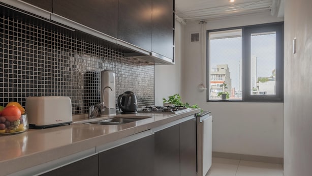 A modern kitchen with dark cabinets, a tiled backsplash, a toaster, a kettle, and a window with a view of buildings. - MOT LIVE HOTEL (Buenos Aires)