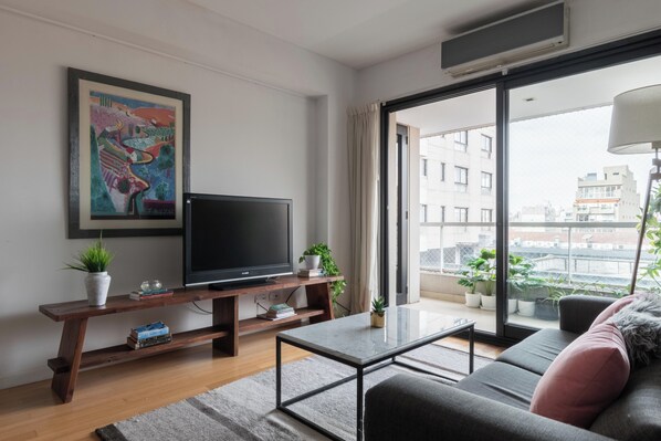Una sala de estar moderna con televisor de pantalla plana, un sofá, una mesa de centro y un balcón con plantas en macetas.