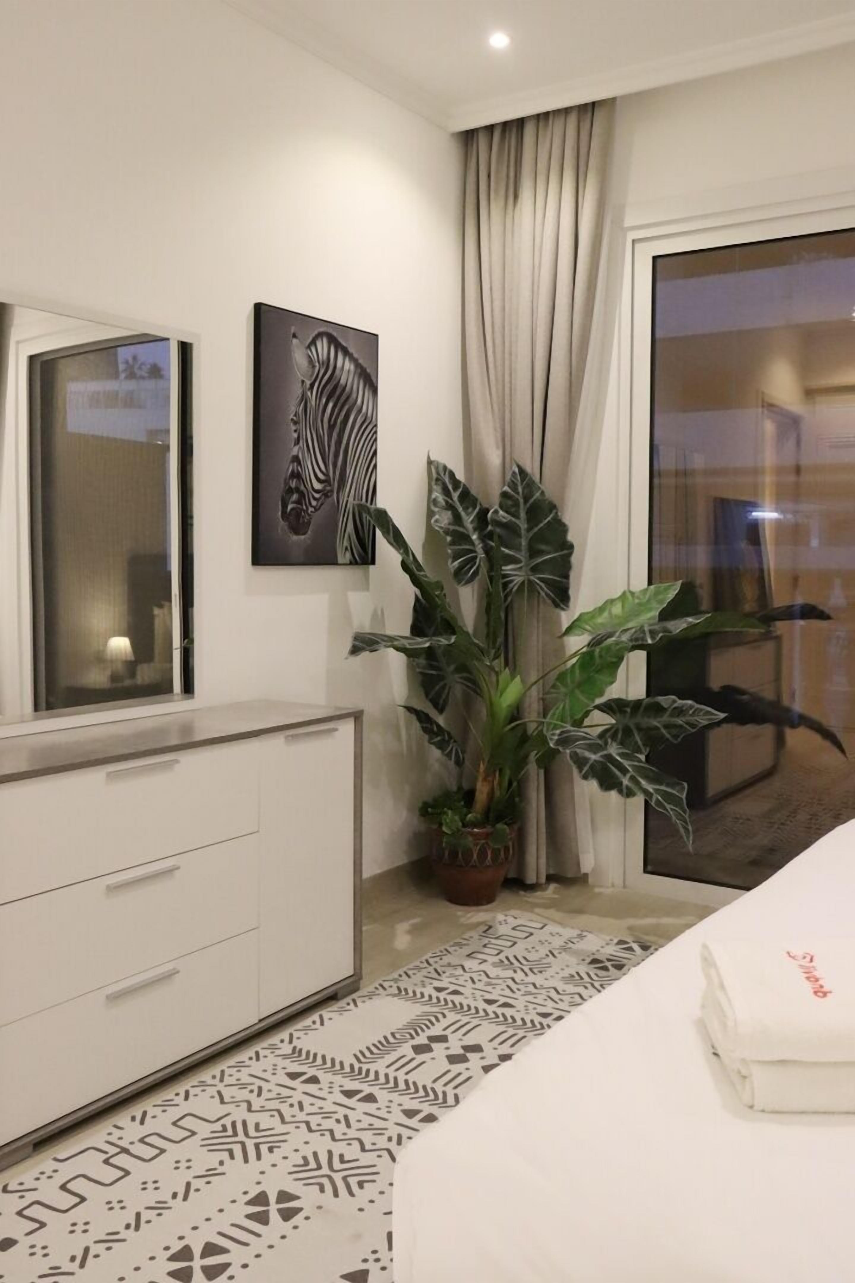Habitación de hotel con cama, cómoda, una obra de arte de cebra, planta grande y vista a través de un ventanal.