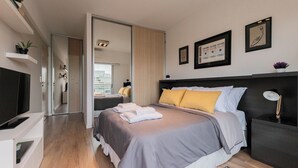 Una habitación de hotel moderna con una cama grande, un televisor de pantalla plana, una mesita de noche con lámpara y vistas a la ciudad desde la ventana.
