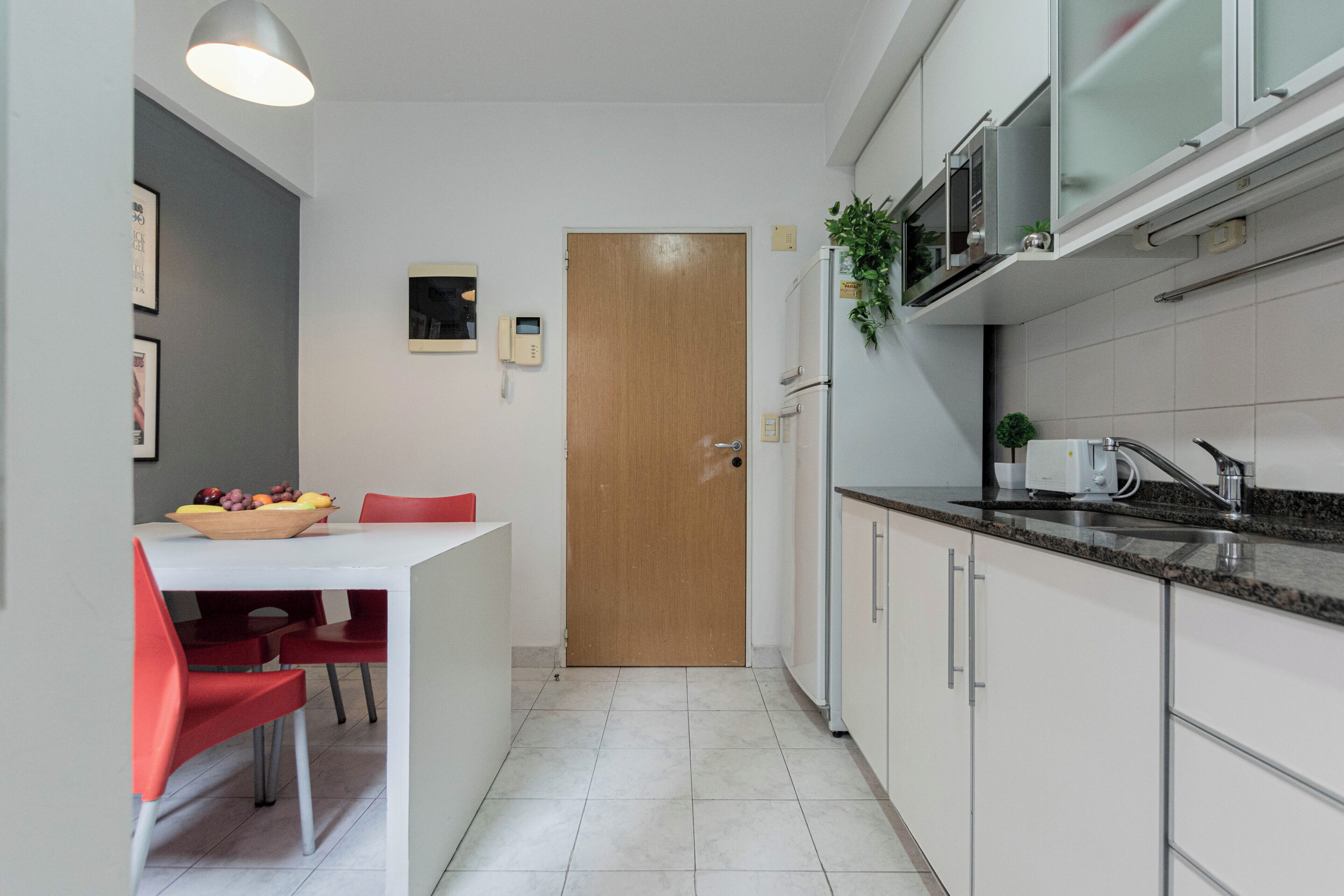 Cocina compacta con armarios blancos, una puerta de madera, una encimera con una cafetera y un frutero sobre la mesa.