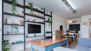 Una sala de estar moderna con una mesa de centro de madera, un sofá azul y un televisor montado en una pared blanca. Se ven estanterías con libros y objetos decorativos.