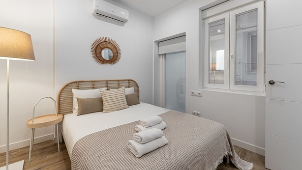 A bedroom with a bed, a bedside lamp, a wall-mounted air conditioner, a mirror, and a window with a door. - Waou Coosy Retiro (Madrid)