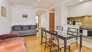 A modern living room with a grey sofa, a red armchair, and a glass dining table with four chairs. A kitchen with white cabinets and a wooden countertop is visible in the background. - Quiet Prince - City Center Apartment (2D) (Lisboa)