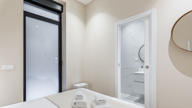 A hotel room with a bed, a bathroom with a glass door, and a round mirror on the wall.