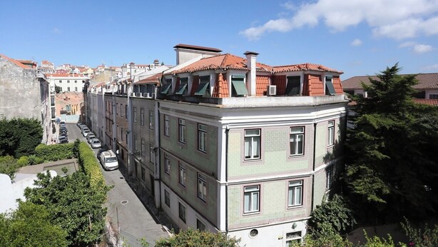 Un edificio de varios pisos con techo de tejas rojas, fachada verde y una calle estrecha en su parte frontal.