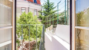 Un balcón con barandilla de metal, una pared blanca y vistas a un edificio de techo rojo y árboles.