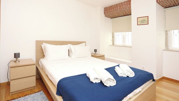 Un dormitorio con cama, mesita de noche, lámpara, ventana y estante de pared.