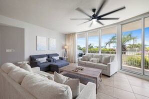 Ein modernes Wohnzimmer mit Deckenventilator, zwei Sofas, einem Couchtisch und einer großen Schiebeglastür, die zu einem Außenbereich mit Blick auf Palmen und einem klaren Himmel führt.
