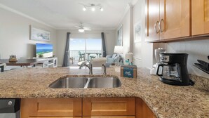 Eine Küche mit Granit Arbeitsplatten, einer doppelten Spüle, Holzschränken, einer Kaffeemaschine und Meerblick durch große Fenster.