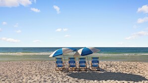 Une plage avec quatre chaises de plage et parasols bleus et blancs, un rivage sablonneux et l’océan en arrière-plan.