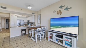 Un salon avec un téléviseur diffusant une scène de plage, un coin repas avec des chaises blanches et une cuisine avec des meubles blancs.
