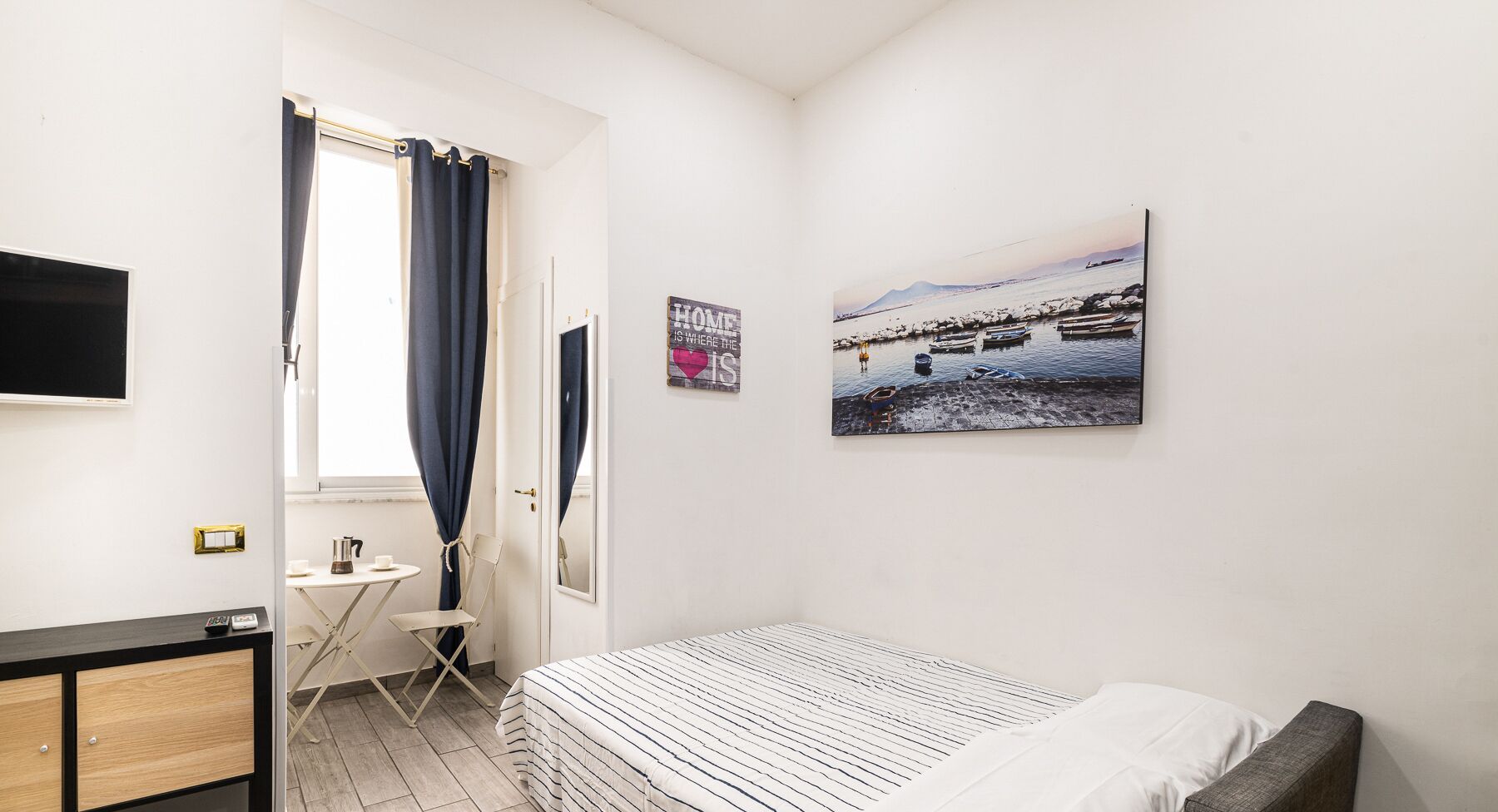 A bedroom with a bed, a small table, a chair, a TV, and a painting of a beach scene.