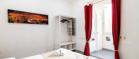 Une chambre d’hôtel avec un lit, une armoire, un rideau rouge, une porte et un tableau représentant un paysage urbain.