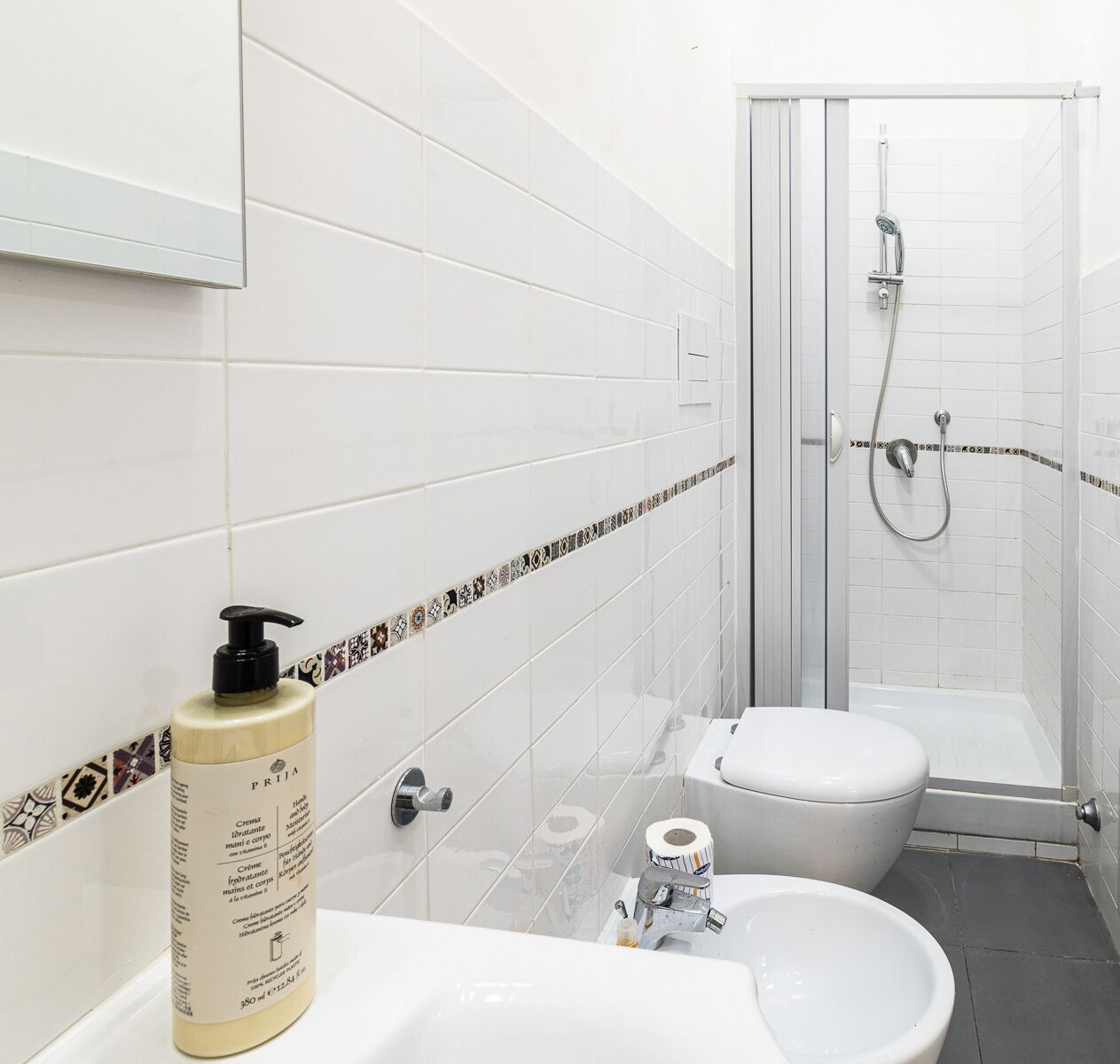 Een badkamer met witte tegels, een toilet, een wastafel, een douche en een flesje lotion.