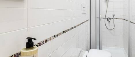 Een badkamer met witte tegels, een toilet, een wastafel, een douche en een flesje lotion.