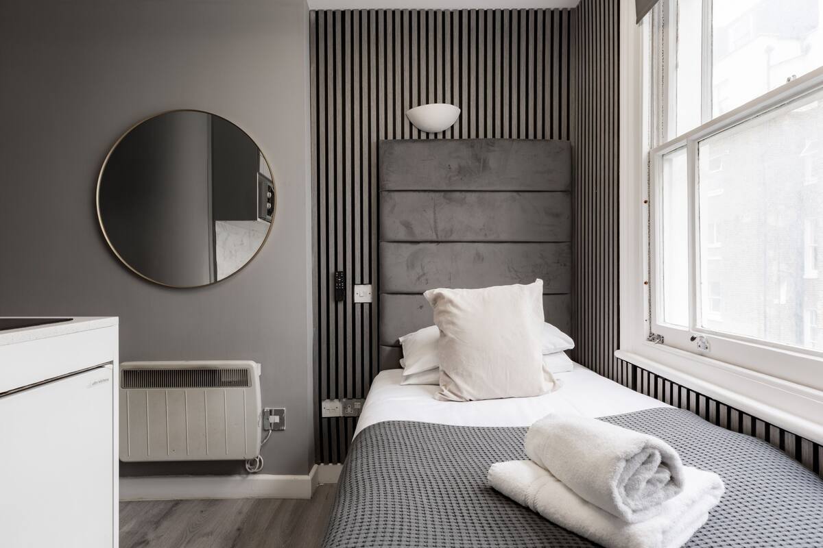 Una habitación de hotel moderna con una cama, un espejo redondo, un radiador y una ventana con vistas.
