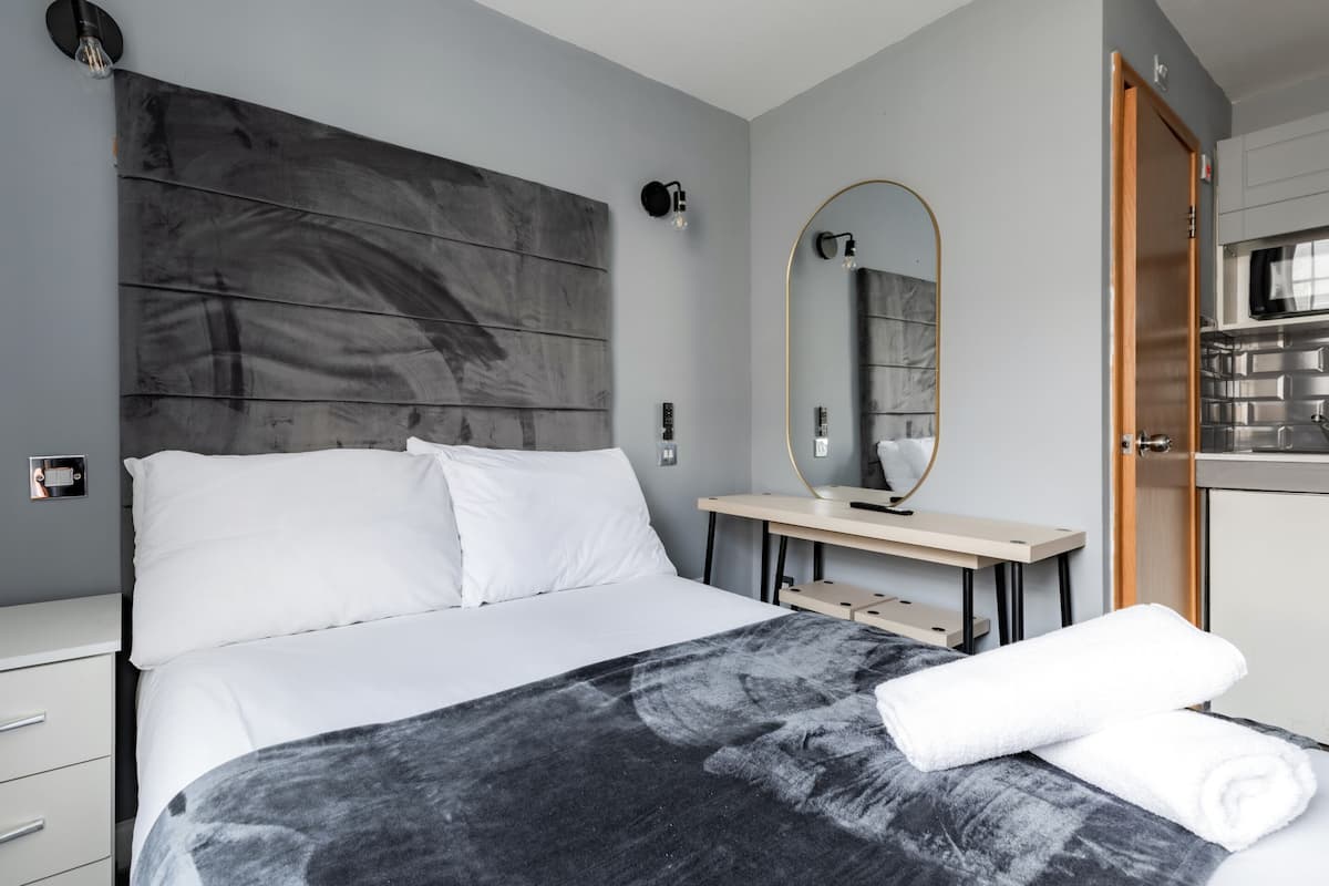 Habitación de hotel moderna con una cama grande, un cabecero de madera, una mesita de noche, un espejo y vistas a la cocina a través de una puerta abierta.