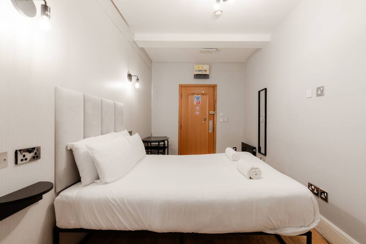Una habitación de hotel con cama, una mesita, una silla, una puerta de madera y un espejo en la pared.