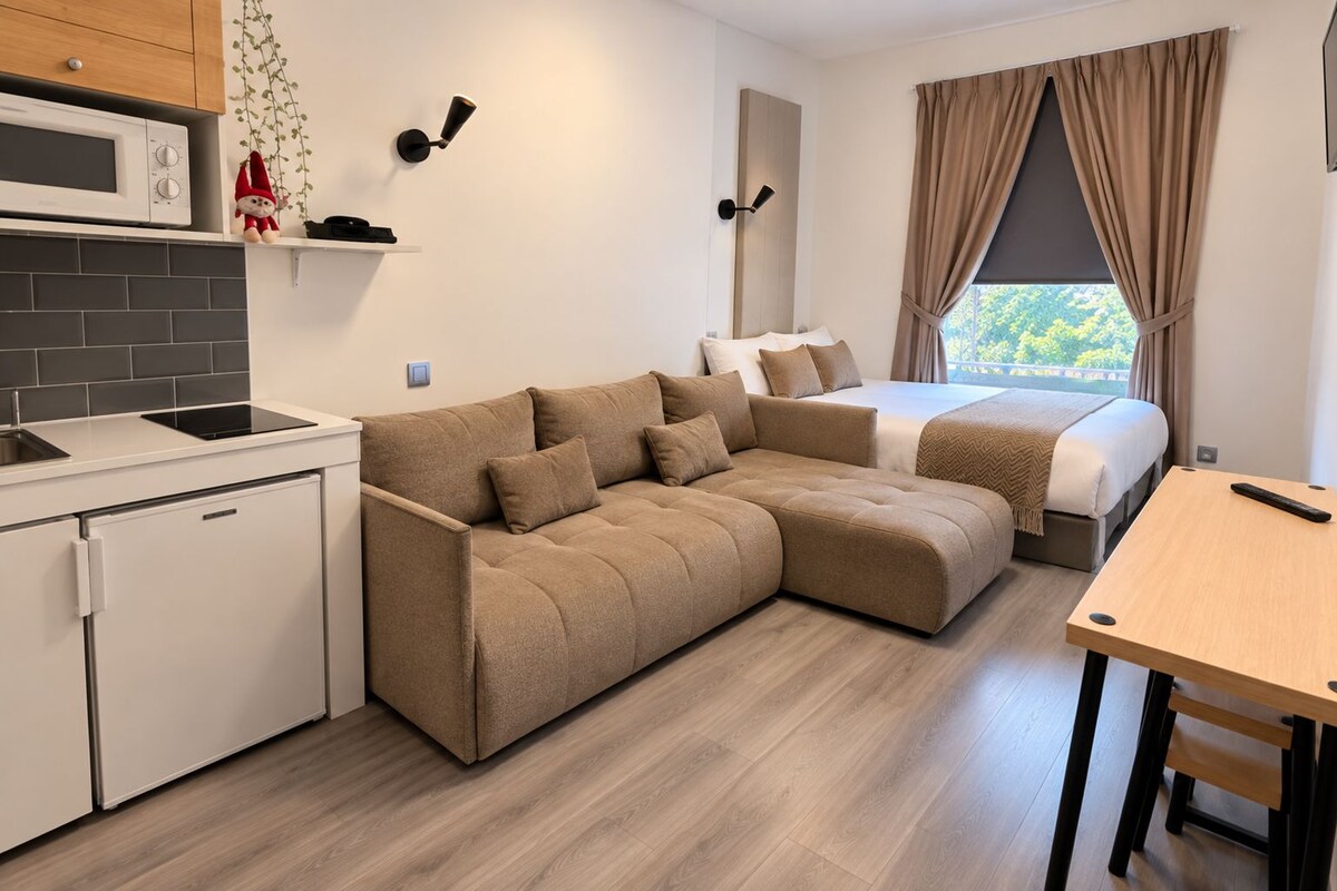 A modern kitchenette with a microwave, a small refrigerator, and a sink. There is a beige sofa and a bed with a view of greenery outside.
