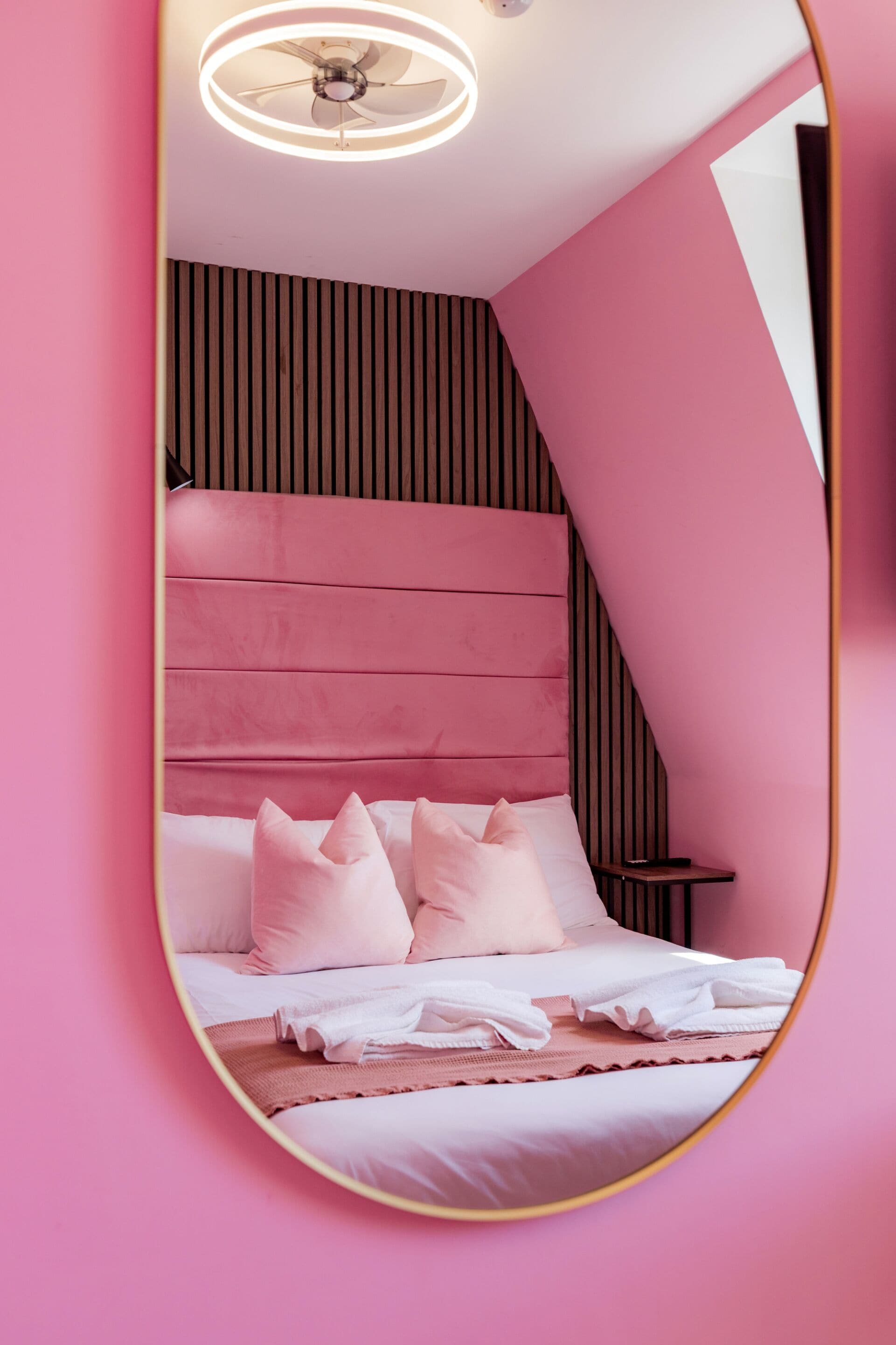 A bedroom with a pink theme, featuring a bed with white linens, multiple pillows, and a ceiling fan.