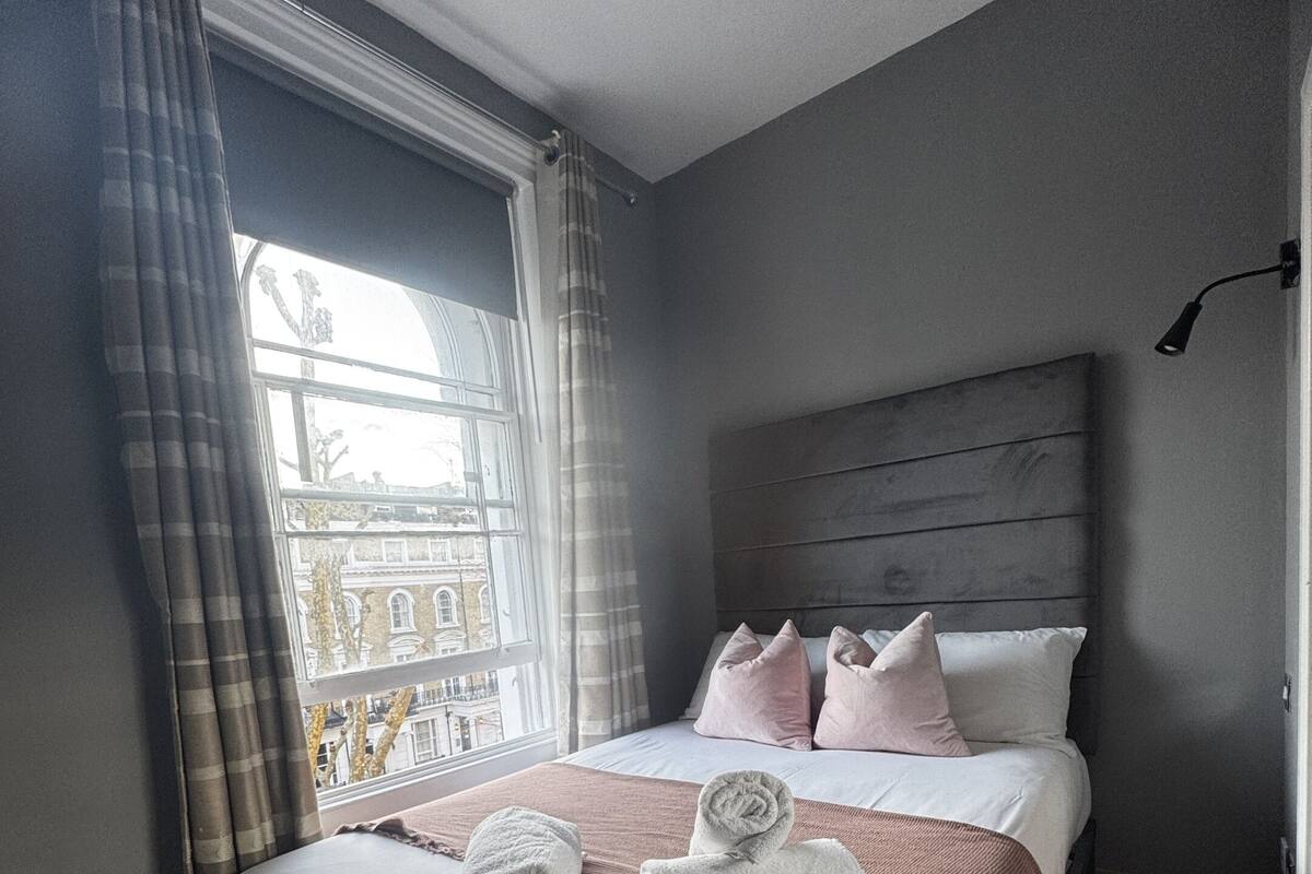 Un dormitorio con un ventanal grande, una cama con almohadas rosa y blancas, un banco y vistas de edificios exteriores.