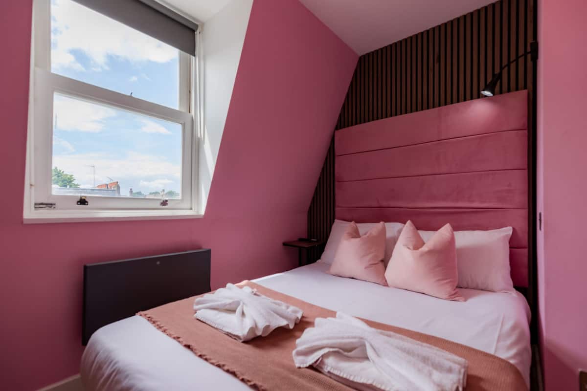 Un dormitorio con cama, almohadas, una mesita de noche y una ventana.