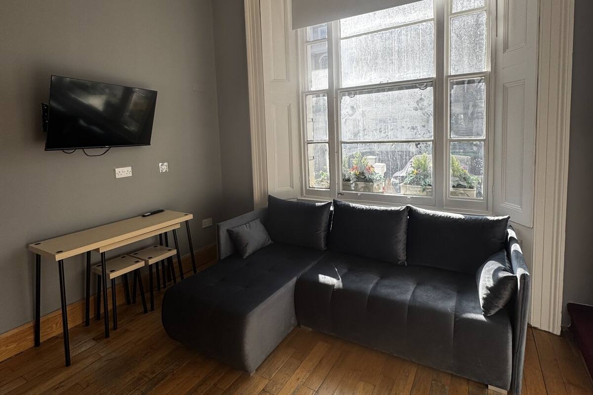 Una sala moderna con un sofá modular, un televisor de pantalla plana en la pared, una mesita de madera y un amplio ventanal con vistas al exterior.