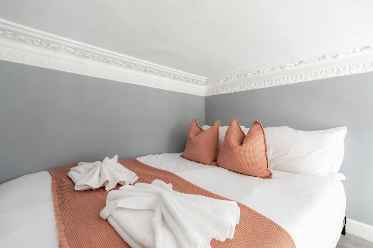 Una cama bien tendida con almohadas blancas y melocotón, una manta color melocotón y una toalla blanca sobre una pared gris.