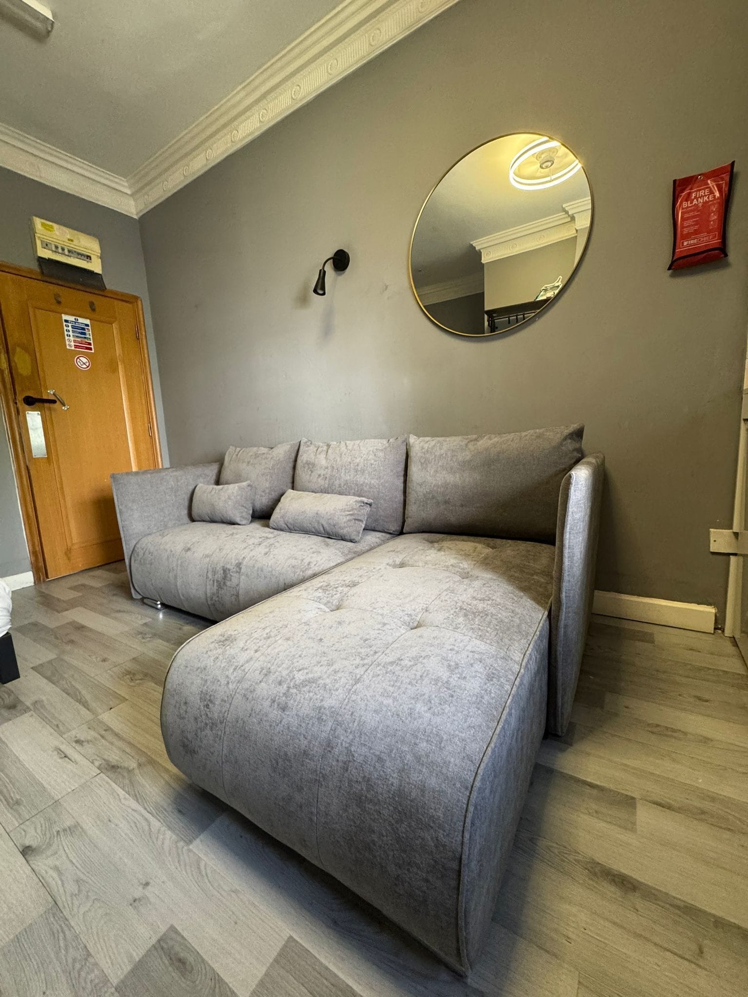 A room with a grey sofa, a round mirror, a wooden door, and a fire blanket on the wall.