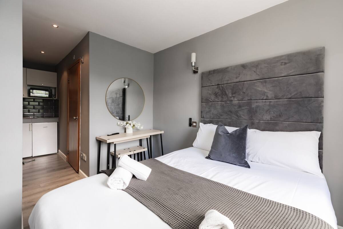 Habitación de hotel moderna con una cama grande, un escritorio pequeño, un espejo redondo y un cabecero gris.