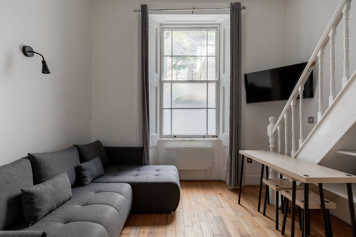 Una sala moderna con un sofá seccional gris, piso de madera, un radiador blanco, un televisor montado en la pared y una escalera con barandales blancos.